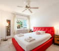 Modern bedroom with a red upholstered headboard bed, white linens, decorative pillows, nightstand with lamp, and a view through large windows.