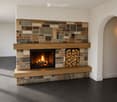 A modern fireplace with a stone surround and wooden mantel, featuring a burning fire and a stack of firewood to the right.