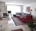 Bright living room with large windows, grey sectional sofa, red armchair, coffee table, fireplace, TV, floor lamp, and decorative items.