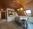 Dining area with wooden ceiling, dining table with chairs, chandelier, mirror, and small kitchen area visible in the background.