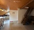 Open-plan living and dining area with wooden ceiling beams, staircase leading to upper level, modern kitchen area with white cabinets and stainless steel appliances, dining table with chairs, and a cozy fireplace area.