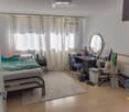 A well-lit bedroom with a bed, desk, chair, mirror, nightstand, and various personal items. The room features wooden flooring and large windows with curtains.