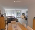 Bright living and dining area with a sofa, dining table, chairs, cabinets, and a television. Large windows with white curtains lead to a balcony.
