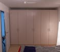 Bedroom with a large built-in wardrobe, a neatly made bed with pillows, and a wooden floor. An open door leads to another room.
