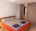 A small bedroom with a wooden bed, orange mattress, grey duvet, and two blue pillows. A white chair is positioned near the window with brown curtains.