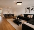 Modern living room with a black sectional sofa, coffee table, dining area with chairs, pendant lights, and artwork on the walls.
