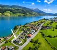Aerial view of a small village nestled by a lake, surrounded by green hills and mountains. The village features numerous houses with varied architectural styles, roads, and a calm lake reflecting the surrounding landscape.