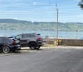 Two parked cars on a paved road near a waterfront with a scenic view of a town and hills across the water.