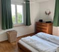 Bedroom with wooden floor, two beds, green curtains, wooden dresser, wicker basket, and a lamp