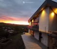 Modern two-story house with a balcony, illuminated exterior lighting, and a scenic sunset view over a rural landscape.
