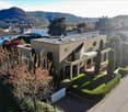 Modern two-story house with a flat roof, large windows, and a spacious garden with neatly trimmed hedges and trees. The house is surrounded by other residential buildings and features a scenic mountain view in the background.