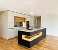 Modern kitchen with white and black cabinets, a central island with a marble countertop, a sink, and an oven. Wooden flooring and recessed lighting.