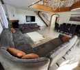 Spacious living and dining area with a large sectional sofa, coffee table, dining table with chairs, television, and a fireplace. The room features a staircase leading to an upper level and large windows with curtains.