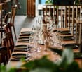 A restaurant setting with a long wooden table set for dining, featuring plates, cutlery, glasses, and a vase with dried flowers.