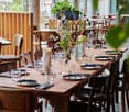 A restaurant setting with wooden tables and chairs, wine glasses, plates, cutlery, candles, and a flower arrangement on the table.