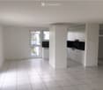 Empty room with white tiled flooring, white walls, and large windows. The space includes a small kitchen area with white cabinets and a countertop.