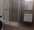 Modern bathroom with a glass-enclosed shower, a white sink with a mirrored cabinet, and a washing machine. The walls are tiled in neutral tones, and the floor is covered with brown tiles.