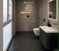 Modern bathroom with dark tiled floor, white sink, toilet, large mirror with LED lighting, and a glass-enclosed shower area with a built-in shelf.
