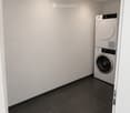A small, modern laundry room with a black tiled floor, white walls, and a white washing machine positioned against the wall.