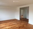 Empty room with wooden herringbone flooring and white walls, featuring an open doorway leading to another room.