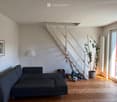 Living room with a grey sectional sofa, wooden floor, white walls, a staircase leading to an upper level, a large potted plant near the sliding glass door, and a floor lamp. A picture frame hangs on the wall, and toys are scattered near the staircase.
