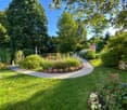 Well-maintained garden with a winding gravel path, lush green grass, colorful flowers, shrubs, and tall trees providing shade.