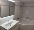 Modern bathroom with a white sink, chrome faucet, bathtub, and a large mirror with built-in lighting. The walls are tiled in a light marble pattern.