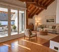 Bright living room with wooden floors, exposed wooden beams, large glass doors opening to a balcony, sofa, coffee table, armchair, side table, floor lamp, and decorative plants.