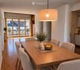 Dining area with a wooden table, six chairs, a vase with greenery, a bowl of fruit, and a candle. The room has wooden flooring, white walls, and a view of a balcony with outdoor seating.