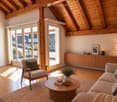 Modern living room with wooden beams, large windows, and a balcony. Features a sofa, armchair, coffee table, and a wooden cabinet with decorative items.