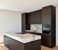 Modern kitchen with a large island, marble countertops, dark wood cabinetry, and built-in appliances including an oven and sink.