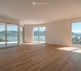 Bright, empty room with wooden flooring and large glass sliding doors leading to a balcony with scenic mountain and lake views.