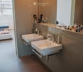 Modern bathroom with two white sinks, mirrors, and various toiletries on the counter. The room features tiled walls and a large window letting in natural light.
