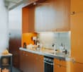 Modern kitchen with wooden cabinets, stainless steel sink, and various appliances. Countertop has a bowl, a water bottle, and other small items. A small table with a ceramic pot and bananas is visible in the foreground.