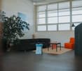 Modern living room with large windows, black sofa, orange chair, small table, and decorative stools.