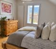 Cozy attic bedroom with wooden flooring, large window offering a scenic view, a wooden bed with a decorative bedspread and pillows, a wooden dresser with a plant, and a framed picture on the wall.