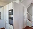 A modern hallway with white walls, wooden flooring, and a staircase leading upwards. The hallway features built-in white cabinets with stainless steel appliances, including ovens and a refrigerator, visible on the left side.