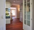 Spacious hallway with wooden flooring, white walls, and recessed lighting. Features a built-in oven and shelves in the background, with a view into an adjacent room with large windows.