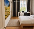 Modern bedroom with a wooden bed, white bedding, a desk with a laptop, a chair, a table lamp, a potted plant, and a large picture frame on the wall. The room features wooden flooring, a large window with dark curtains, and a textured rug.