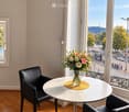 A small, modern room with wooden flooring, a round white table, two black leather chairs, and a vase of flowers on the table. Large windows offer a scenic view of the city and water.
