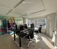 Modern office space with large windows, sleek black desk, ergonomic chairs, and multiple computer monitors.