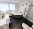 Modern bathroom with a white sink, bathtub, toilet, and glass-enclosed shower. Features include a large window with blinds, white tiles, and a dark tiled floor.