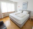 Cozy bedroom with a double bed, white bedding, a grey headboard, a black leather chair, a small wooden stool, a picture frame on the wall, and a floor lamp.