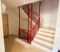 Modern hallway with a staircase featuring red metal railings and white steps, leading to an upper level. The walls are white, and there is a wooden door visible on the left side.