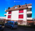 Multi-story residential building with red shutters, white facade, and a parking area with a silver van.