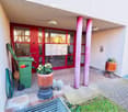 Entrance area with a red double door, potted plants, a green trash bin, and a small garden patch.