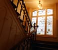 Staircase with ornate wooden railing and stained-glass windows, warm lighting from a chandelier.