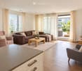 Modern living room with a brown sectional sofa, wooden coffee table, armchairs, TV, and large windows leading to a balcony.