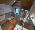 Modern bathroom with wooden flooring, a double sink, toilet, bidet, and a glass-enclosed shower area. Features include a towel rack with a towel and an orange bathroom mat.