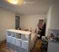A small, cozy bedroom with a bed, storage unit, mirror, clothing rack, and a cart with shoes. The room features wooden flooring and a slanted ceiling with a round light fixture.